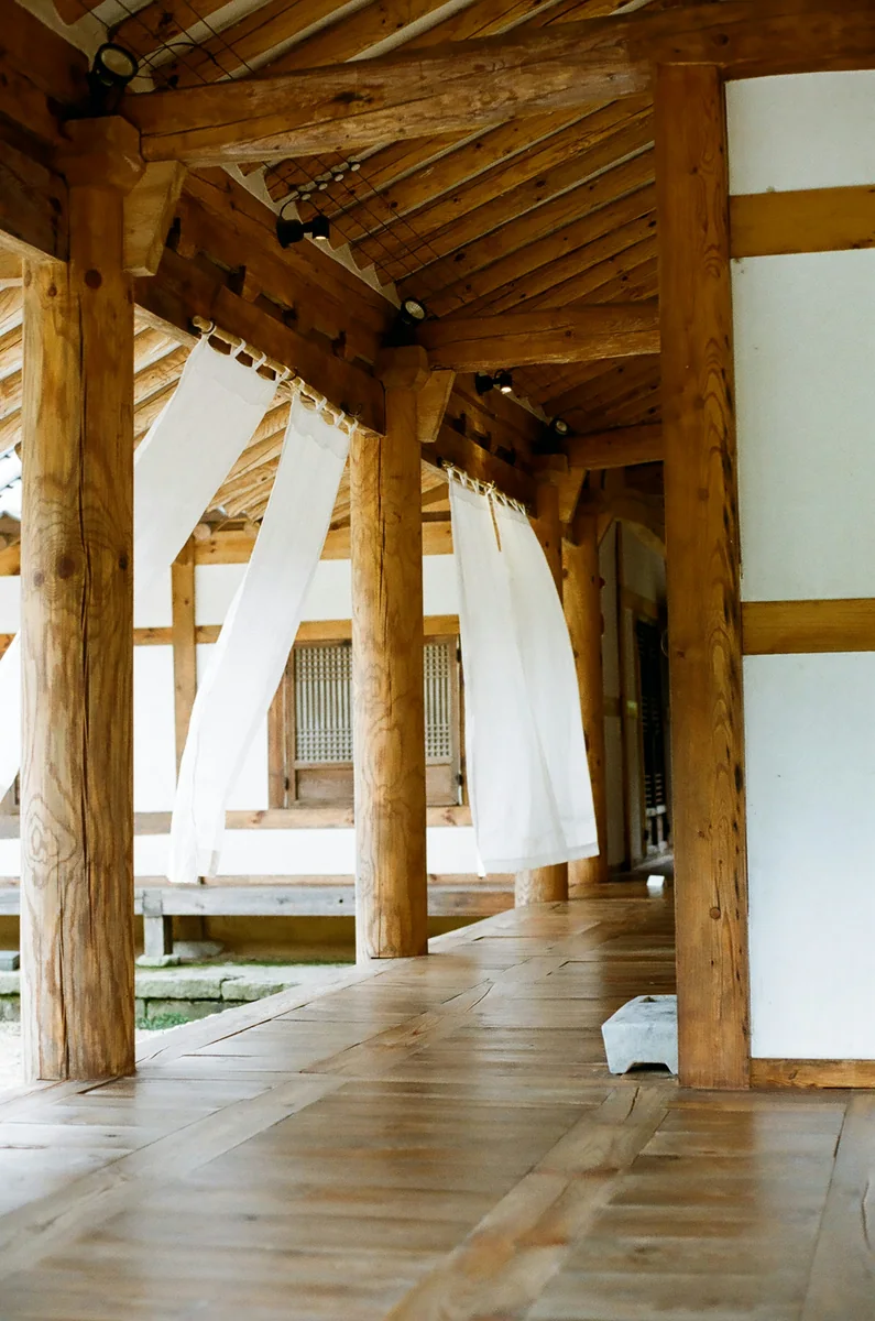 Wooden floorboards and posts beside the doors of a hanok, with white curtains.